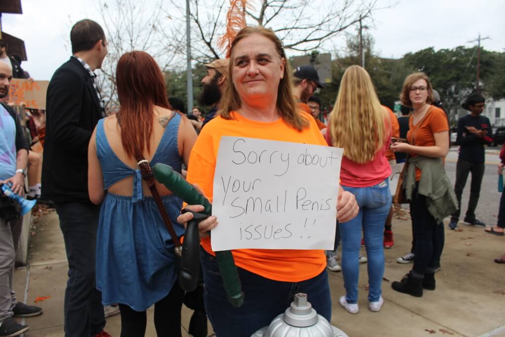 A woman holds three dildos and a sign implying unflattering motivation for those orchestrating a mock mass shooting. (Anna Casey)